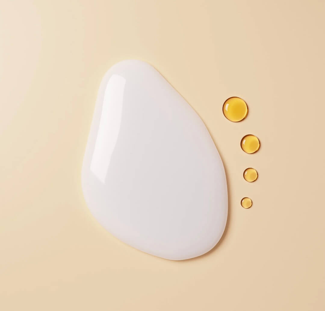 One big white cream drop next to four smaller oil drops on a beige surface.