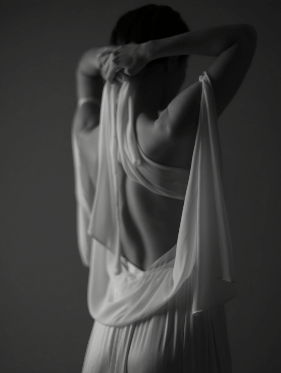 Black and white image of a woman from behind in a white, backless dress with her hands behind her head.