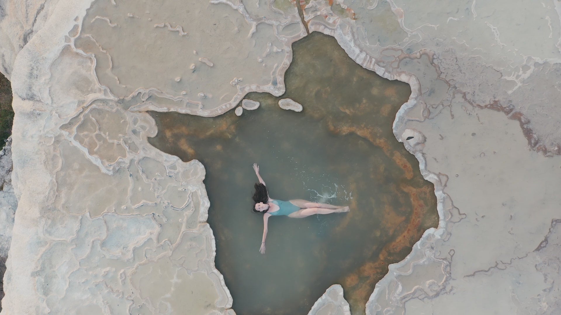 Video of a woman lying in a tide pool next to the ocean. She is wearing a blue bathing suite and floating in the water. It is a birds view shot zooming out.
